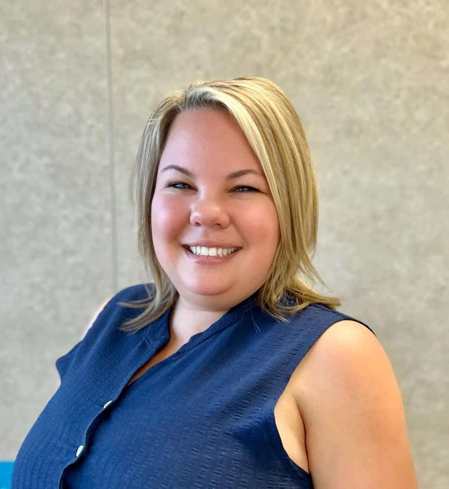 Ashley, a smiling woman in a blue shirt, standing in front of a wall.